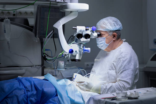 A surgeon in full sterile attire — white gown, gloves, surgical mask, and cap — leans into a surgical microscope while operating on a patient in an operating room. Medical equipment and monitors are visible in the dimly lit background, and the patient is draped in blue surgical cloth.